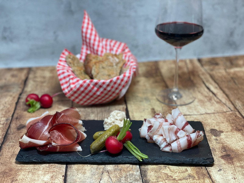Speck aus Südtirol angerichtet auf Schieferplatte mit Radisschen und Gurken. Im Hintergrund steht ein Glas Rotwein und Brot
